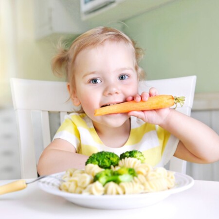 dziewczynka z talerzem makaronu z brokułami je marchewkę